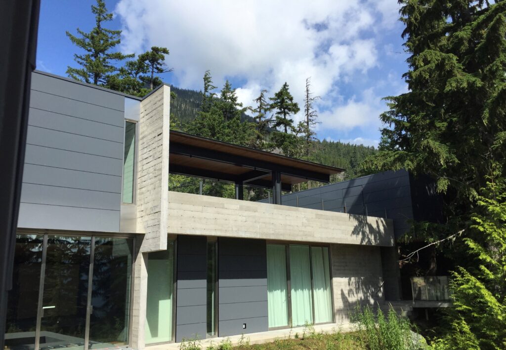 Modern two-story house with concrete and metal exterior, large windows, and surrounded by tall trees and mountains under a partly cloudy sky.