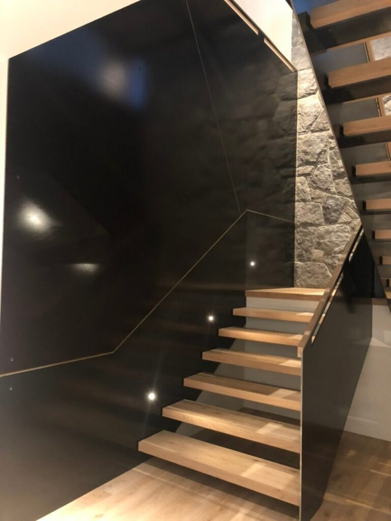A modern staircase with wooden steps, a minimalist black wall, and a stone accent wall on the right. Small lights are embedded along the side of the stairs.