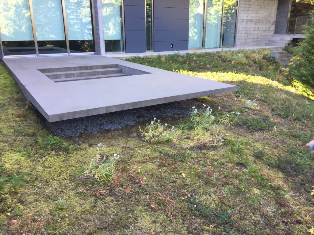 A modern concrete patio with a recessed square feature extends over a grassy area, next to a building with large glass windows and gray paneling.