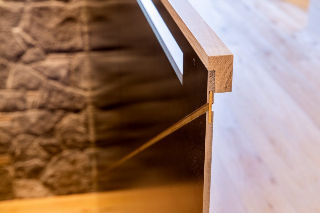 Close-up view of a wooden and glass railing detail with a metal support, next to a stone wall and light wood flooring.
