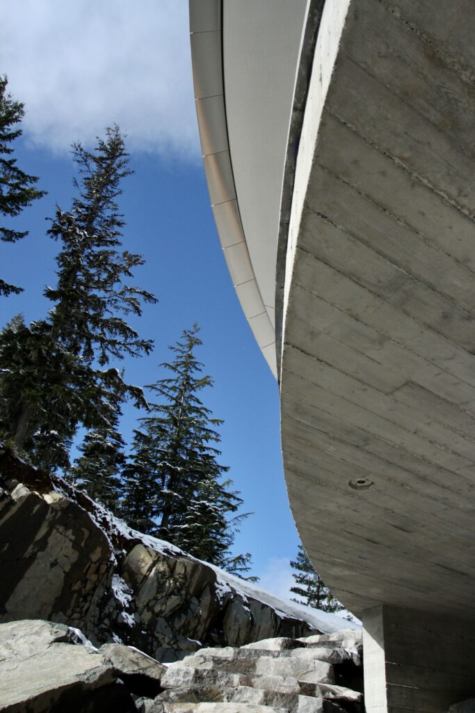 Concrete building overhang viewed from below, with rocks, tall pine trees, and a blue sky with scattered clouds in the background.
