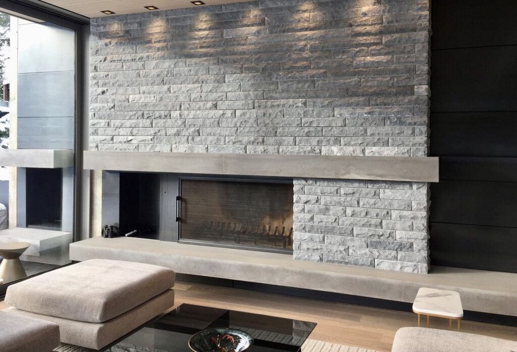 Modern living room with a large gray stone fireplace, built-in concrete mantel, glass coffee table, and neutral-colored seating.