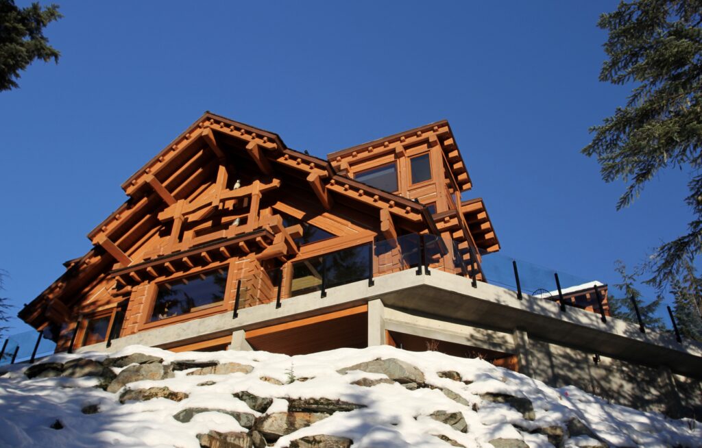 Large wooden log cabin with glass balcony sits on a concrete foundation amid snow and rocks, surrounded by trees under a clear blue sky.