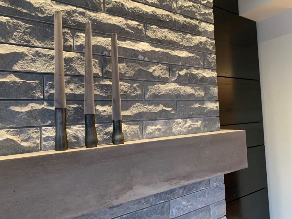 A stone accent wall with a wooden mantel displays three modern decorative candle holders of varying heights.