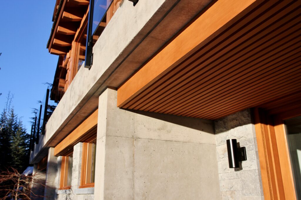 Close-up view of a modern house exterior featuring concrete walls, wood trim, horizontal wood slats, and large glass windows under bright sunlight.
