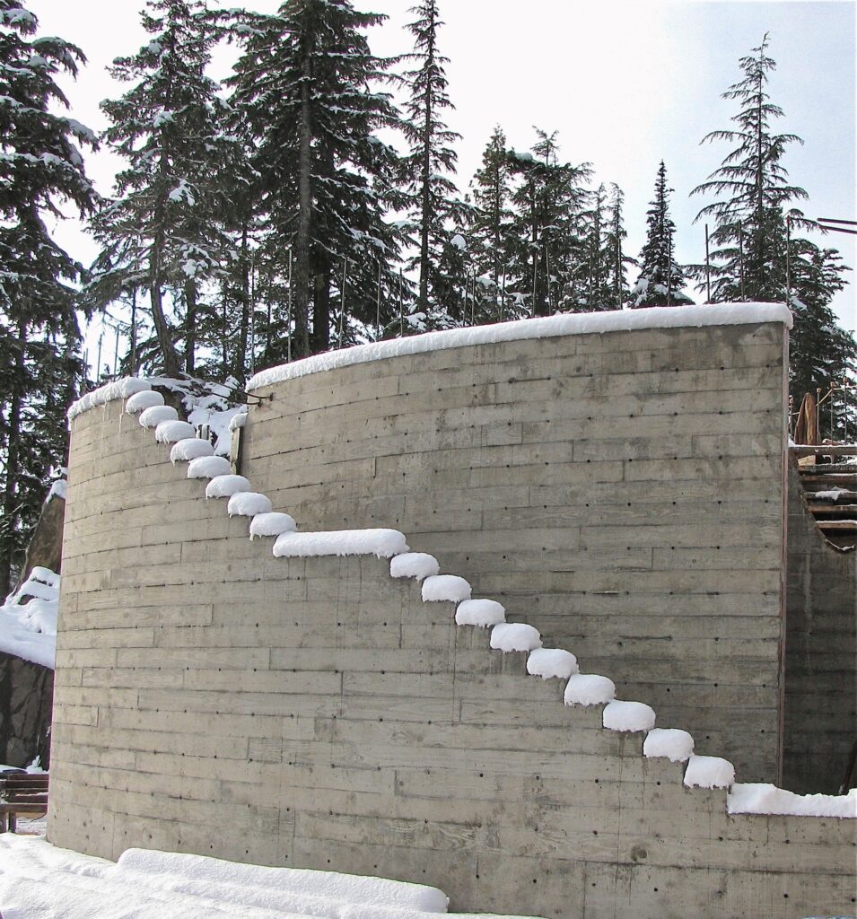 Curved concrete wall with external stairs, each step coated in snow, surrounded by snow and tall evergreen trees.
