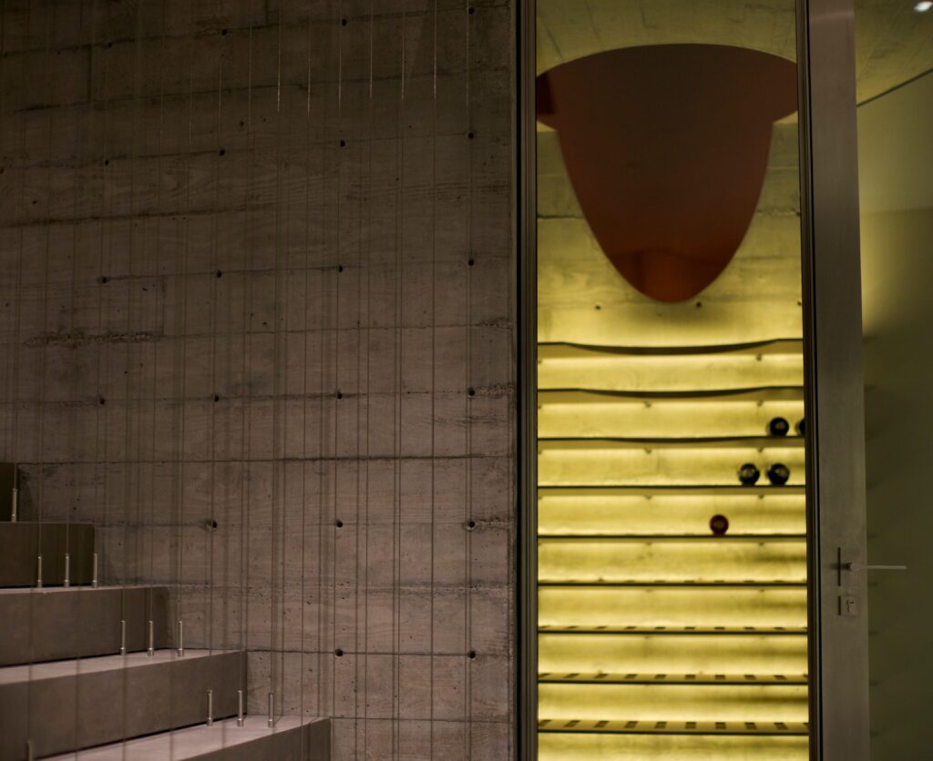 A modern, minimalistic wine cellar with illuminated wooden shelves and several wine bottles, adjacent to a concrete staircase and wall.