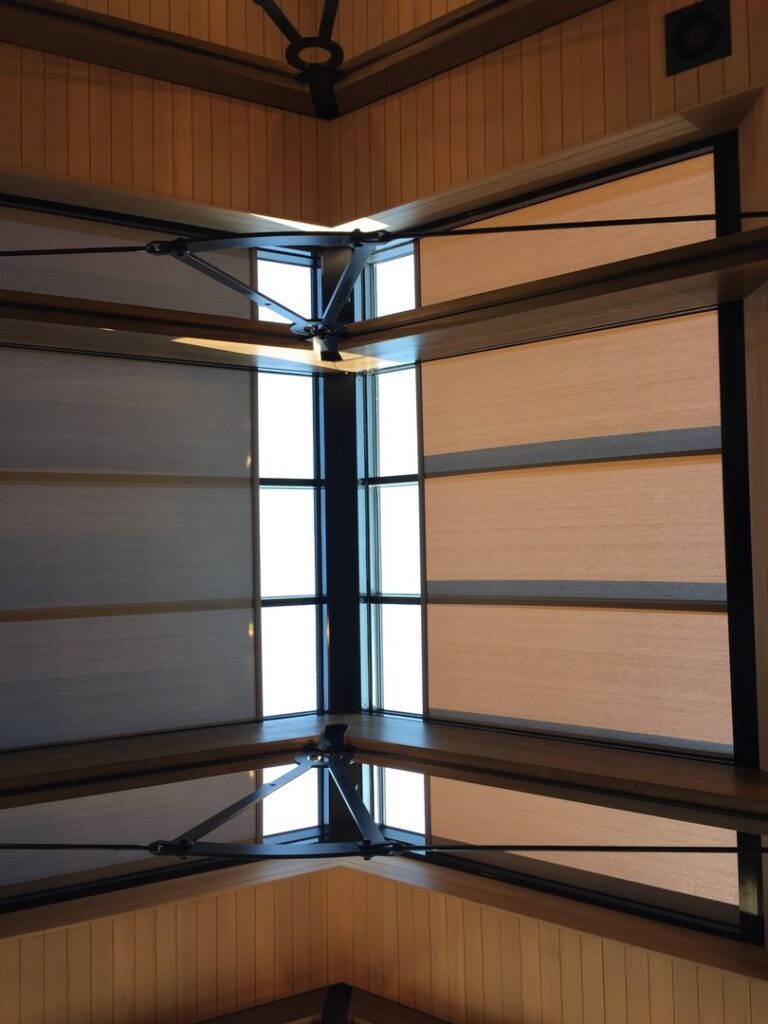 Ceiling view of a room with large windows, metal beams, and partially closed roller shades allowing some natural light in.