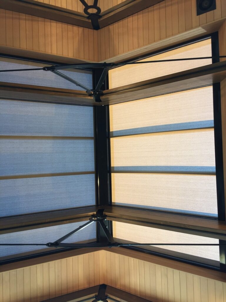 View of a glass ceiling with metal beams and partially open beige and blue fabric shades, framed by light wood paneling.