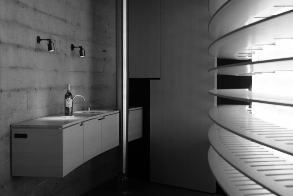 A modern, minimalist kitchen with a wall-mounted sink, a bottle on the counter, concrete walls, and curved shelves on the right. Black and white.
