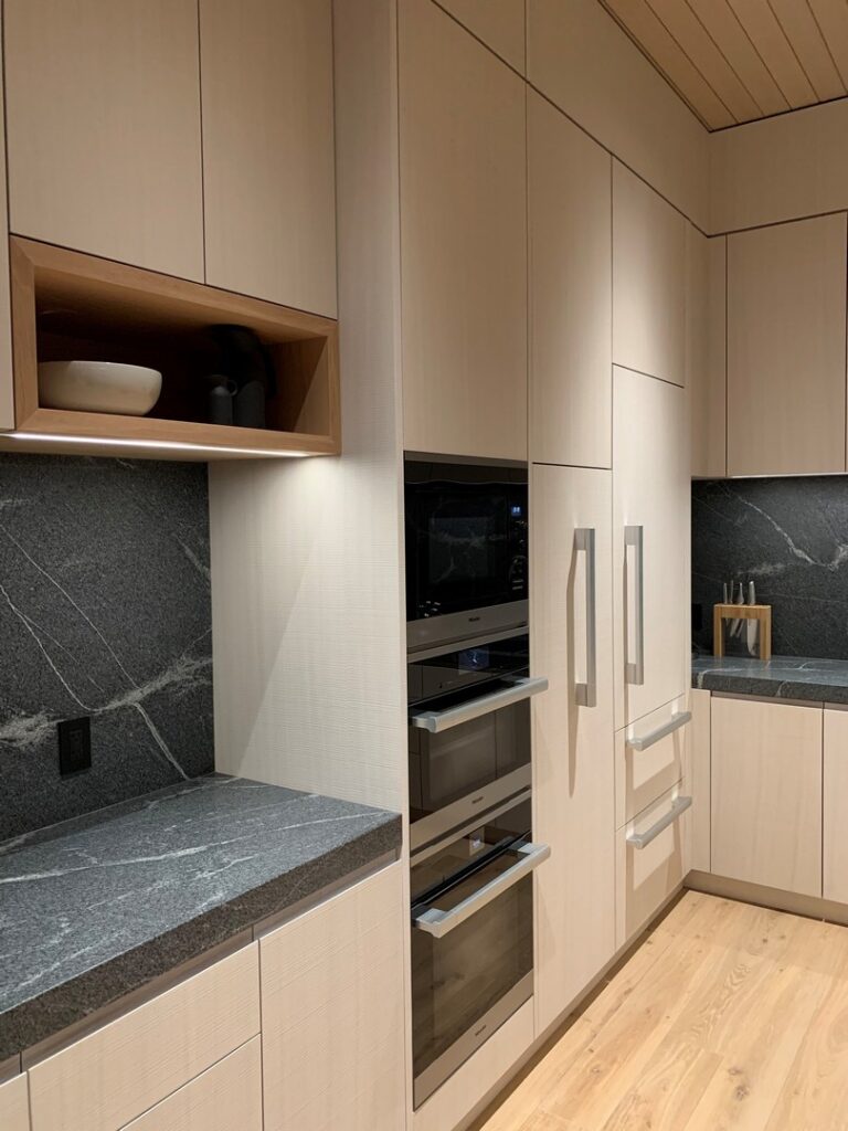 Modern kitchen with light wood cabinets, built-in stainless steel appliances, black stone countertops, and a shelf with bowls and containers.