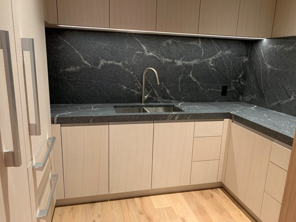 Modern kitchen with light wood cabinets, black stone countertop and backsplash, built-in sink, under-cabinet lighting, and wood flooring.