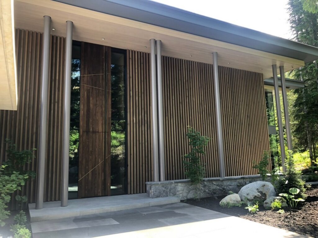 Modern house exterior with vertical wooden slats, tall windows, metal columns, stone steps, and landscaped garden with rocks and plants.