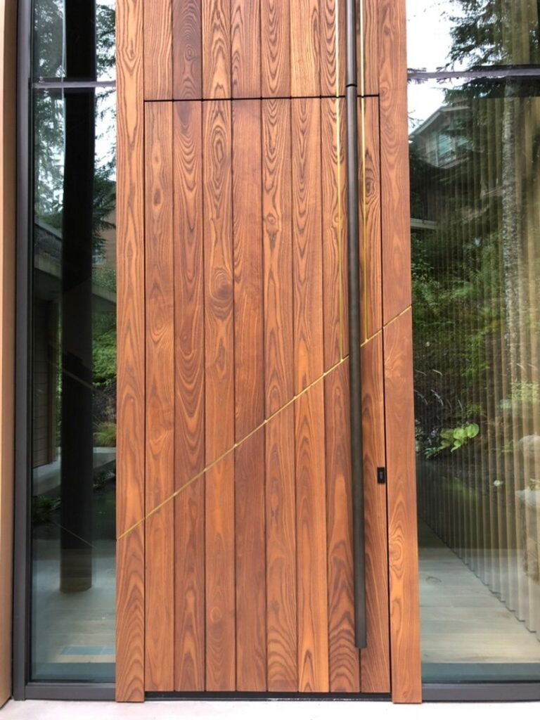 A modern wooden door with vertical wood grain panels, a minimal black handle, a small keyhole, and a diagonal yellow line detail; glass walls reflect trees outside.