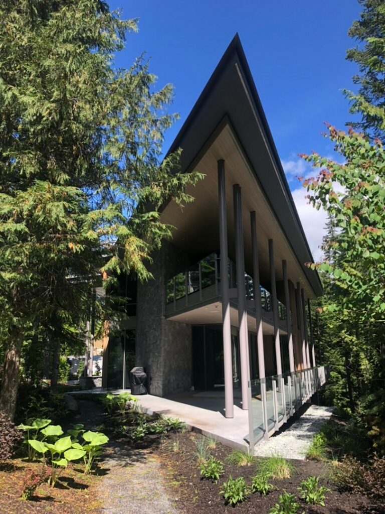 Modern house with tall pillars and a sharply angled roof, surrounded by trees and greenery under a blue sky.
