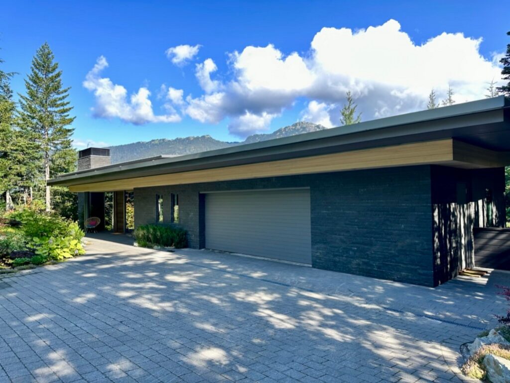 Modern single-story house with a flat roof, dark exterior, and two-car garage, surrounded by trees and mountains under a partly cloudy sky.