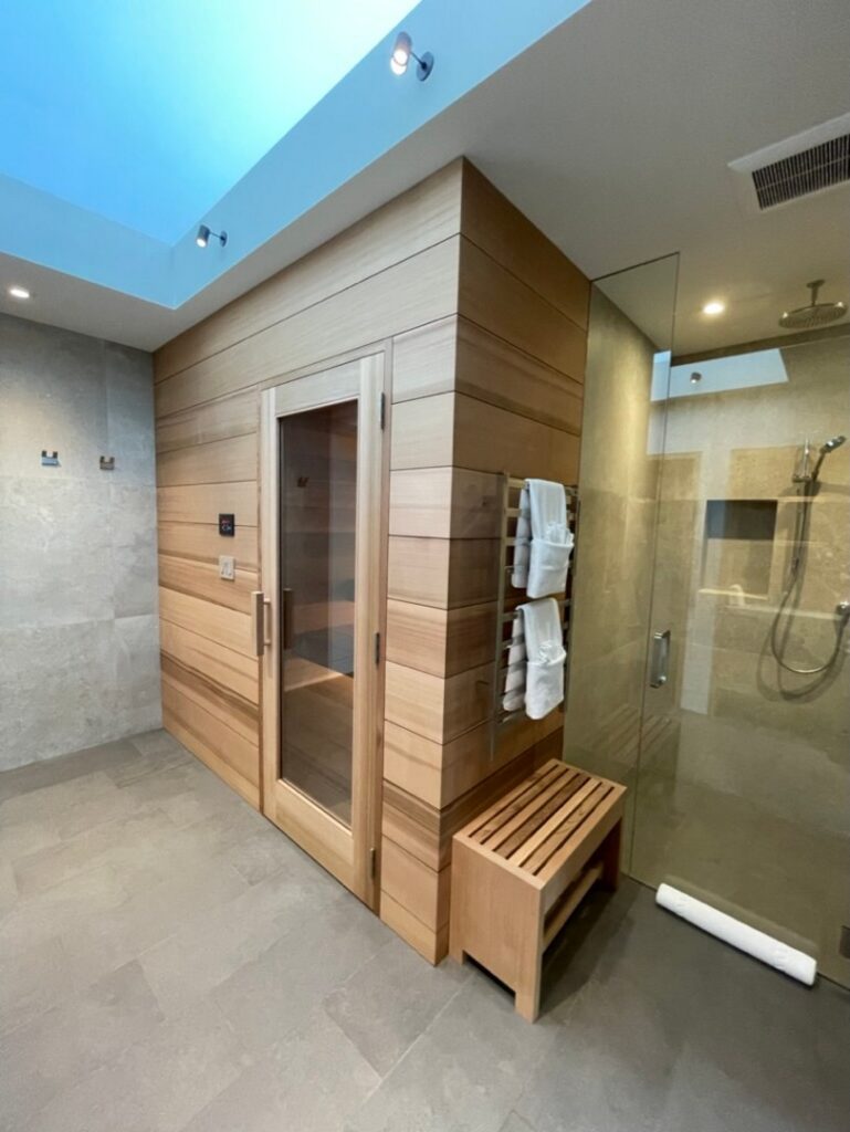 A modern bathroom with a wooden sauna, glass shower area, towel rack, wooden bench, and grey tiled floors and walls.