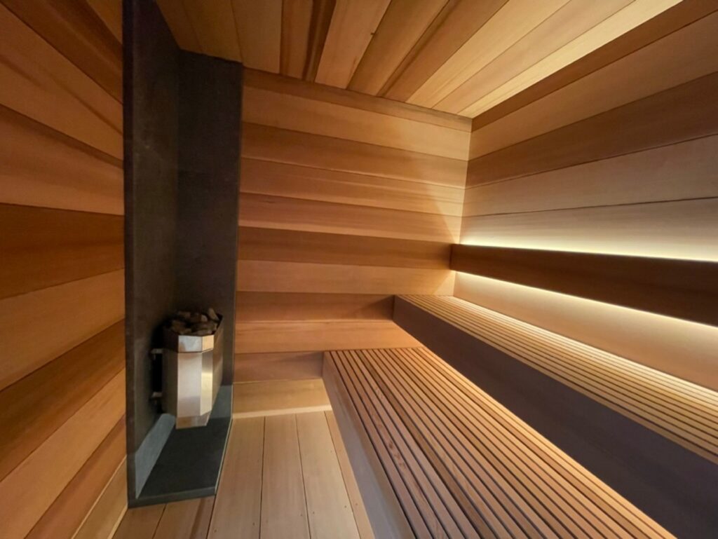 Interior of a modern wooden sauna with tiered seating, a stone heater in the corner, and soft built-in lighting along the bench and wall.