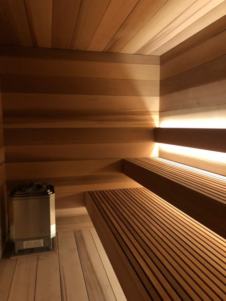 Wood-paneled sauna interior with tiered bench seating and a heater with rocks in the corner, lit by soft wall lights.