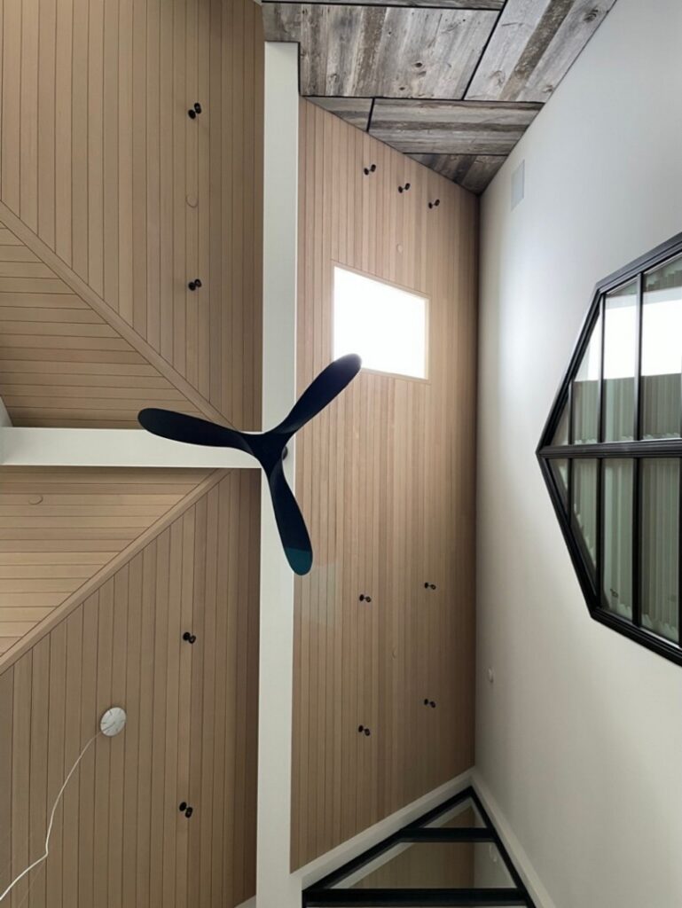 A modern interior with wood-paneled walls, a black three-blade ceiling fan, a square window, and geometric mirrors reflecting the room.