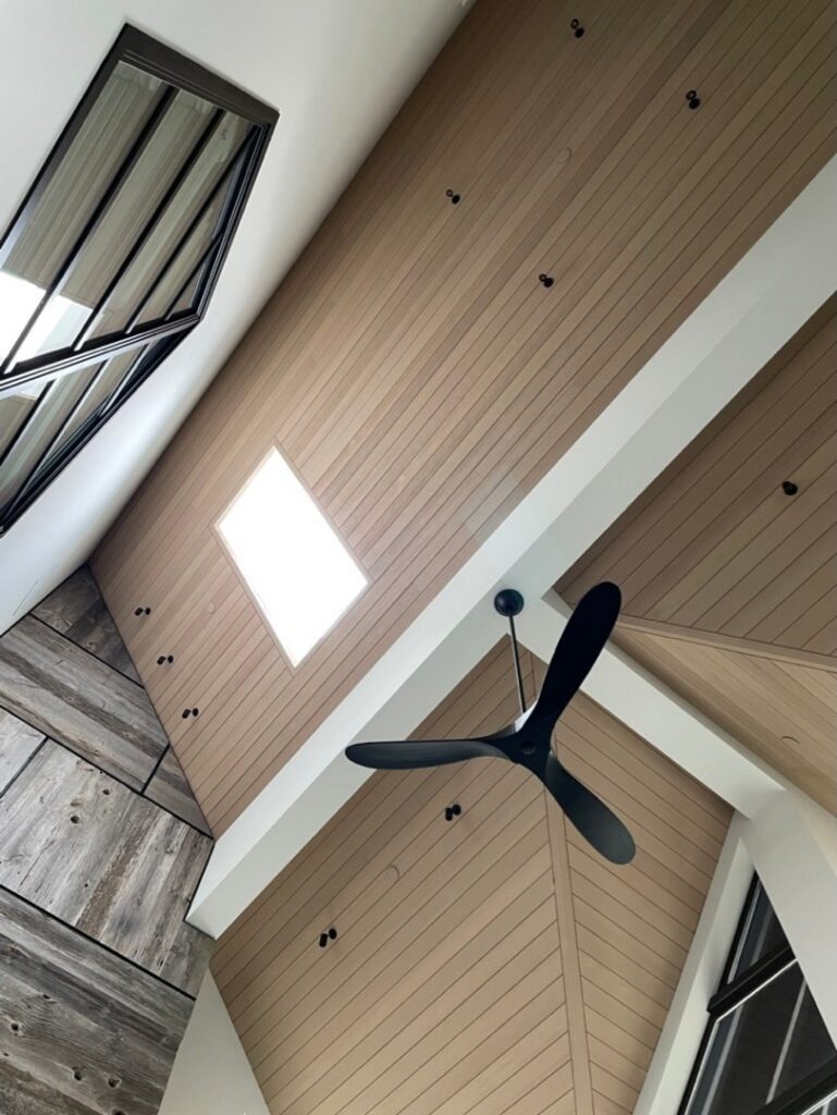 A modern ceiling with light wood paneling, a black ceiling fan, a rectangular skylight, and angled windows with black frames.