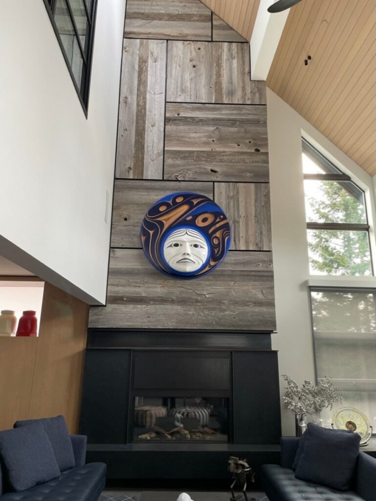 A circular mask with a white face and blue, orange, and black patterns hangs above a fireplace on a tall wooden accent wall in a modern living room.