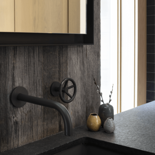 A modern bathroom sink with a matte black wall-mounted faucet, dark countertop, and three small decorative vases next to a wooden-framed mirror.