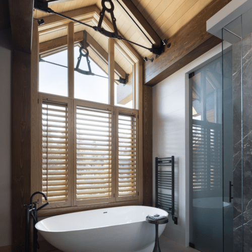 A modern bathroom features a freestanding white tub by large wooden windows with shutters, a glass shower enclosure, and exposed wooden beams on the ceiling.