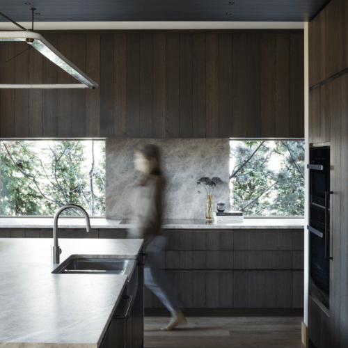 A modern kitchen with dark wood cabinets, marble countertops, two large windows, and a person walking past the island sink.