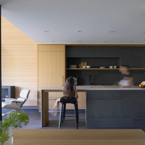 A person sits on a stool at a kitchen island while another person moves nearby; the kitchen features wood and dark cabinetry with minimalist decor.