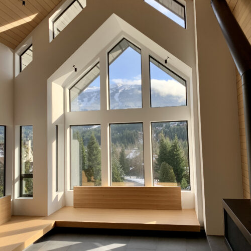 Modern living room with large geometric windows, wood bench seating, and mountain views through the glass. Natural light fills the space with a black wood stove on the right.