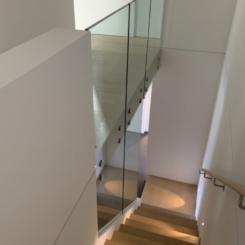 A modern indoor staircase with wooden steps, glass railing, and wall-mounted lights, leading down to a landing with natural light.