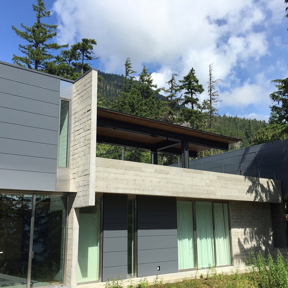 Modern two-story house with concrete and gray metal panels, large windows, and surrounded by trees and mountains under a partly cloudy sky—designed in collaboration with the Custom Building Network.