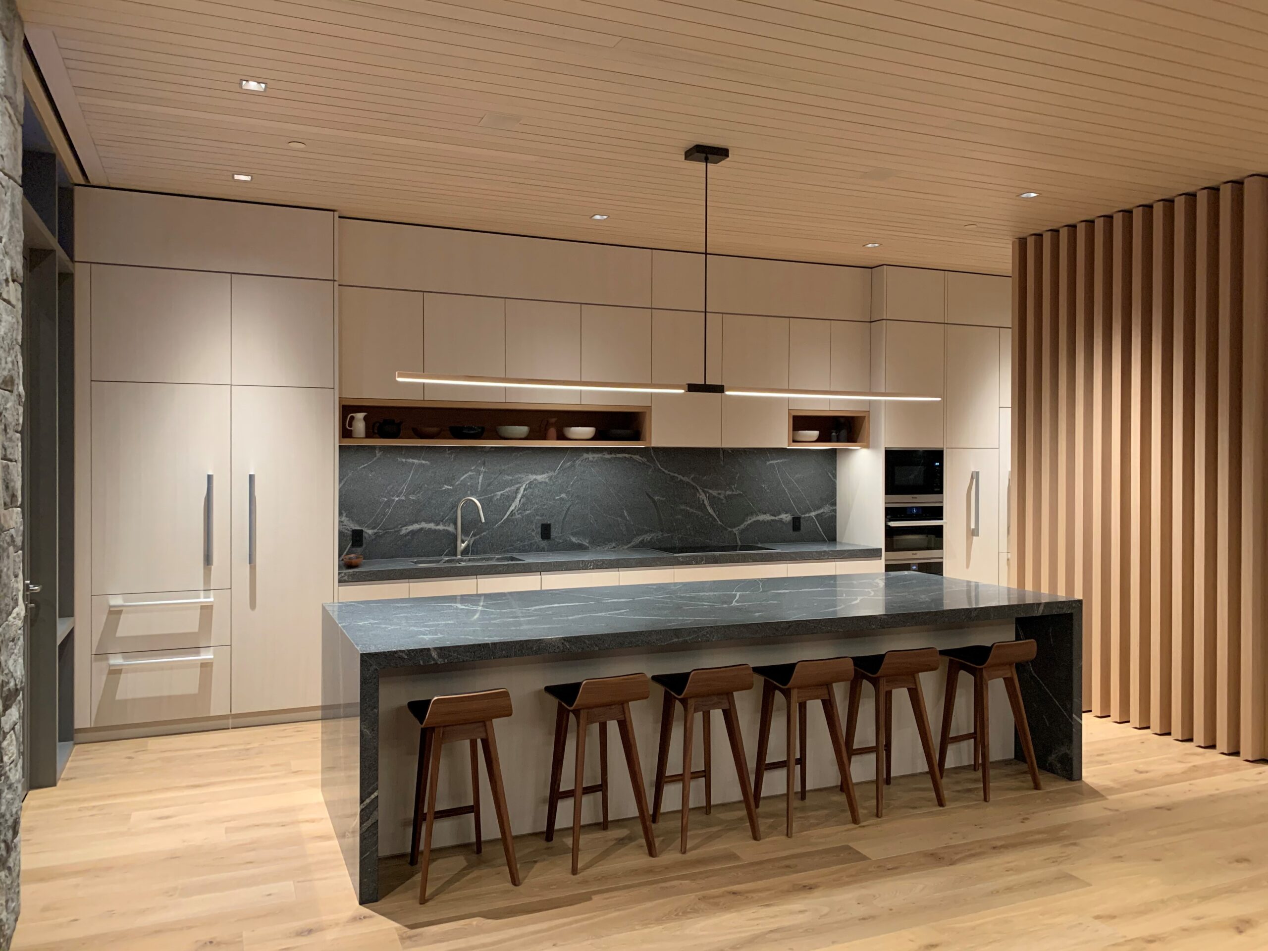 Modern kitchen with light wood cabinetry, a custom-built dark stone island and five wooden stools, black marble backsplash, and minimalist pendant lighting—crafted with care by the Custom Building Network.
