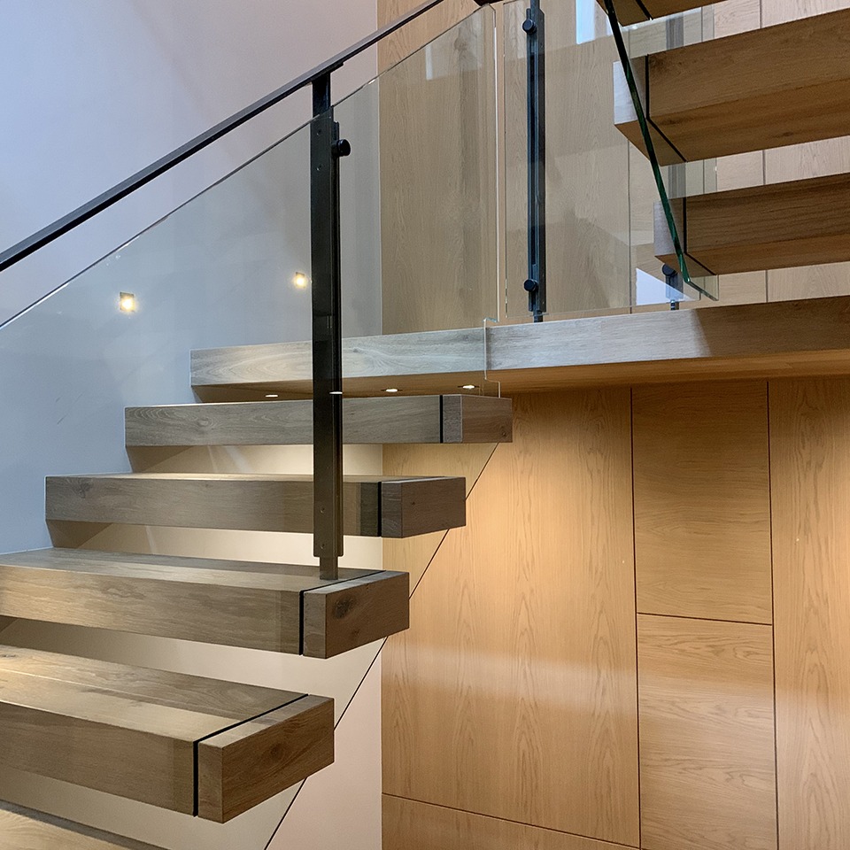 A modern wooden staircase with floating steps, glass railing, and metal supports against a light wood-paneled wall—crafted with precision by the Custom Building Network.